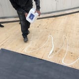 DACHPROTECT Flächenkleber BlueTek wird aus dem Kanister auf eine Holzfläche gegossen, weiße Klebstoffbahnen