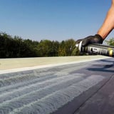 Hand sprüht grünen Flächenkleber Spraybond auf eine schwarze Dachbahn, blauer Himmel und Bäume