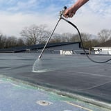 Sprühlanze von DACHPROTECT in Aktion, eine Hand sprueht Fluessigkeit auf eine Dachbahn