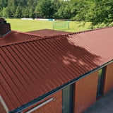 Trapezblech 35/207 rotbraun auf einem Gebaeude mit Schornstein, im Hintergrund ein Sportplatz