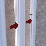 Zevener Sprosse Wandleiste 16 mm, weisses PVC Profil mit Schraubbefestigung an einer Wand, rote Pfeile zeigen auf Schrauben