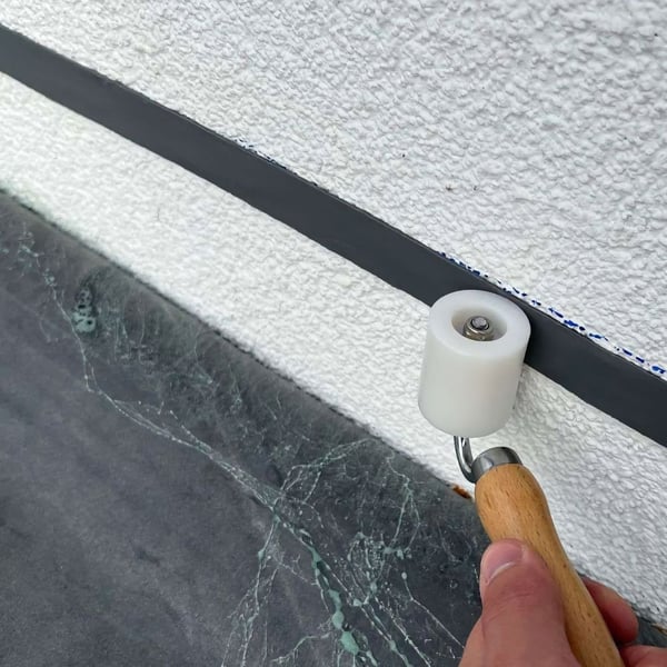 Graues doppelseitiges Dichtband wird mit einer Rolle an einer weissen Wand befestigt
