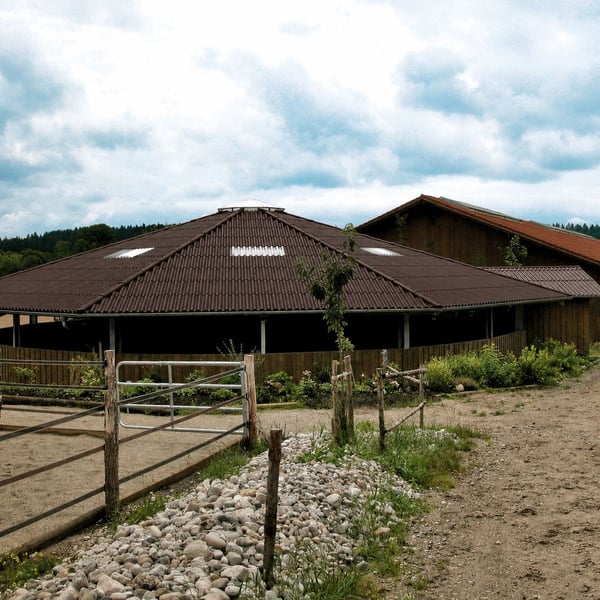 Dunkelbraunes Faserzement Wellplattendach auf einem runden Reitstallgebäude mit Holzzaun im Vordergrund