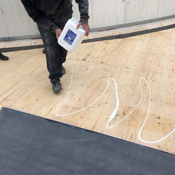 DACHPROTECT BlueTek Flächenkleber wird aus einem weißen Kanister auf eine Holzoberfläche gegossen
