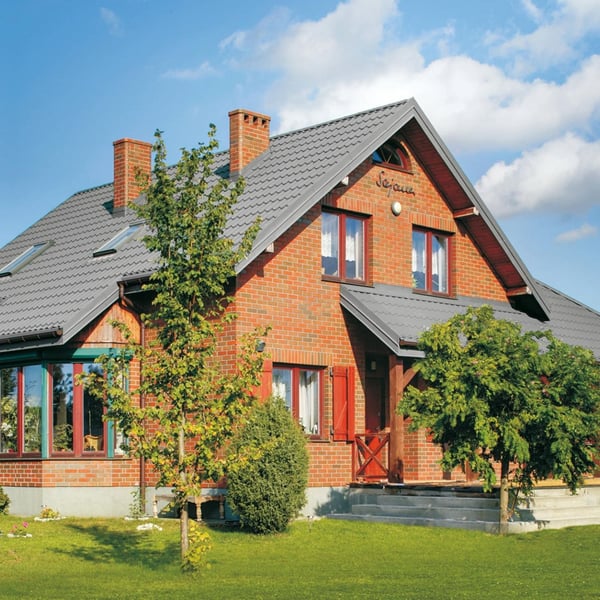 Rotes Backsteinhaus mit Dach aus Pfannenblech Szafir 350/15 in Weißaluminium, umgeben von Bäumen