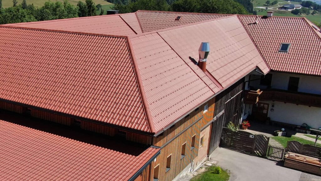 Ländlicher Gebäudekomplex mit großflächigen Dächern aus rot beschichtetem Pfannenblech in Ziegeloptik, kombiniert mit Holzfassaden, gelegen in hügeliger Landschaft mit Ausblick auf grüne Wiesen und Wälder.