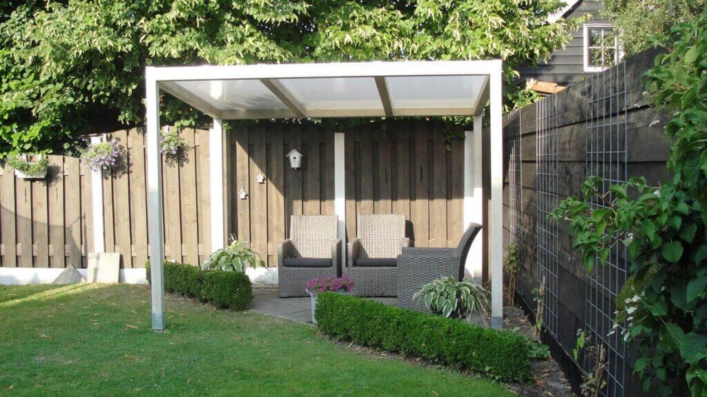 Terrassenüberdachung aus Aluminium mit klarer Kunststoff-Dacheindeckung in einem gepflegten Garten, darunter Sitzgruppe aus Polyrattan, umgeben von Holzzaun und Bepflanzung.