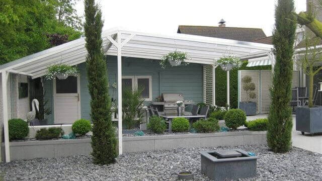 Großzügige Terrassenüberdachung aus weißem Aluminium mit klarer Dacheindeckung, integriert in modern gestalteten Garten mit Zierkies, Hochbeeten, Formschnittgehölzen und Gartenmöbeln vor einem hellblauen Gartenhaus.